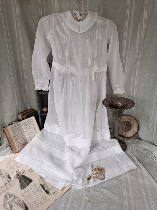 French Brocante - For Sale - c1900 Fine Muslin Child's DressDescription: <p>Dating from the turn of the century this young girl's dress would have been part of a premier communion ensemble, it is made from a light and sheer&nbsp;cotton muslin and is entirely sewn by hand.</p>

<p>The details are beautiful, tiny hand sewn pin tucks on the body, cuffs and skirt hem, drawn thread details, a sweet Pierrot style collar with cloth buttons, tiny lace trim on the cuffs and the highlight an integral belt which finishes at the front with two muslin fabric roses attached to the waist&nbsp;with poppers.</p>

<p>It fastens with a row of cloth covered buttons at the back.</p>

<p>It is in great condition, fully laundered,&nbsp;these dresses are ethereal by nature and need careful wearing but can be worn with a slip or body underneath, chest 30", waist 28", length 38"</p>
